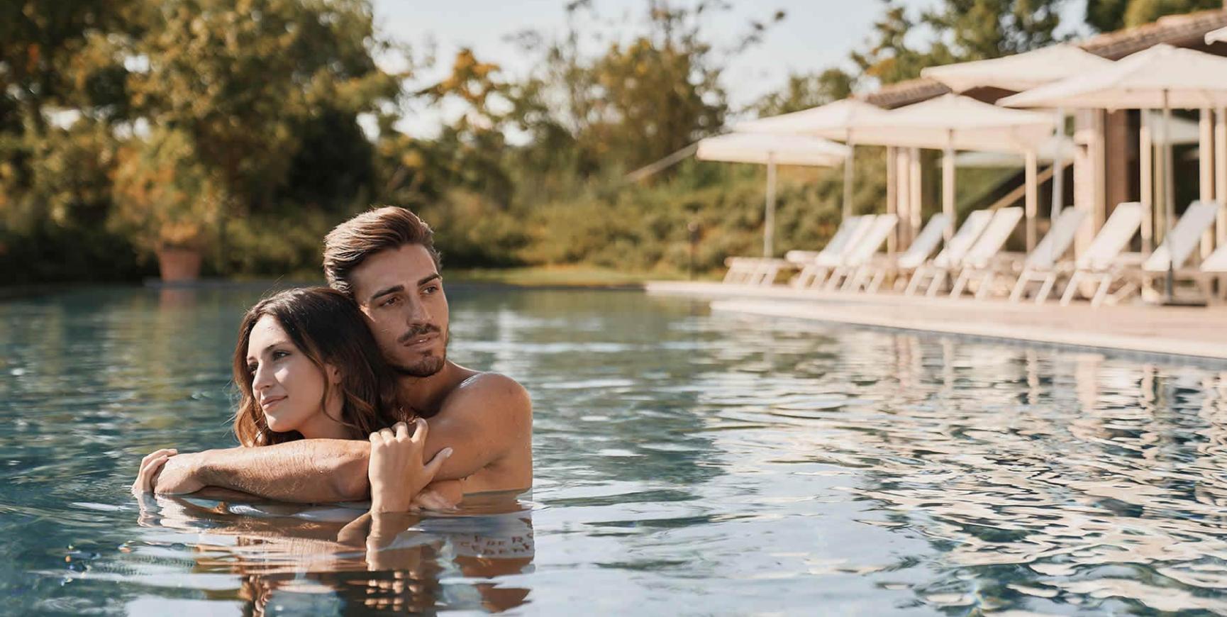 Coppia abbracciata in piscina, circondata da lettini e ombrelloni.