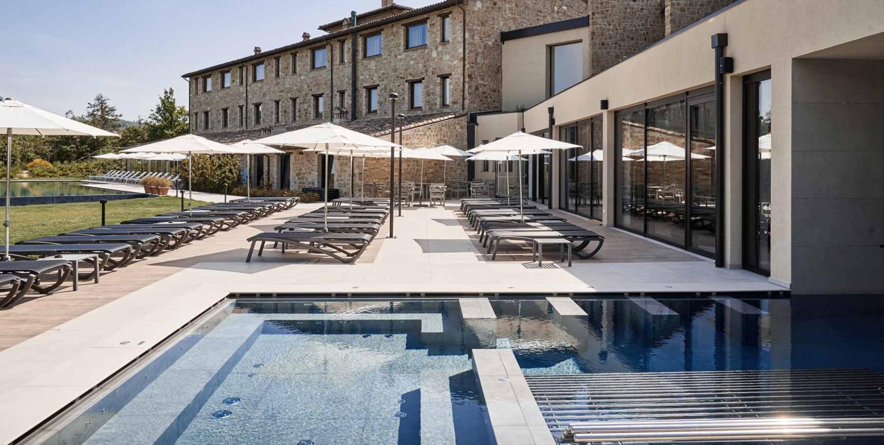 Piscina esterna con lettini e ombrelloni, edificio in pietra sullo sfondo.