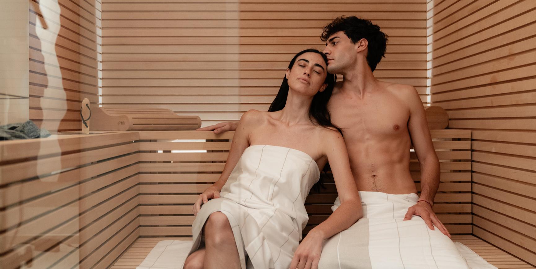Coppia rilassata in sauna, avvolta in asciugamani bianchi.